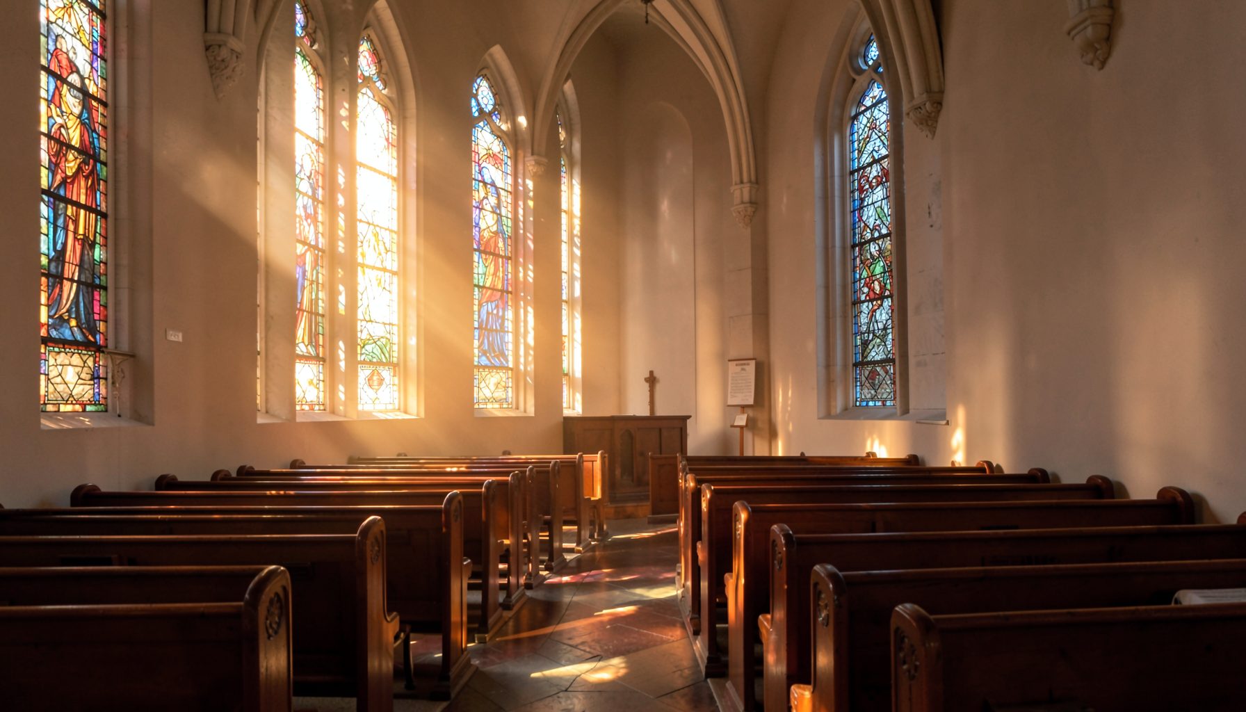 Sunlight filtering through tall stained-glass windows in a quiet sanctuary, casting warm amber and soft blue hues across wooden pews