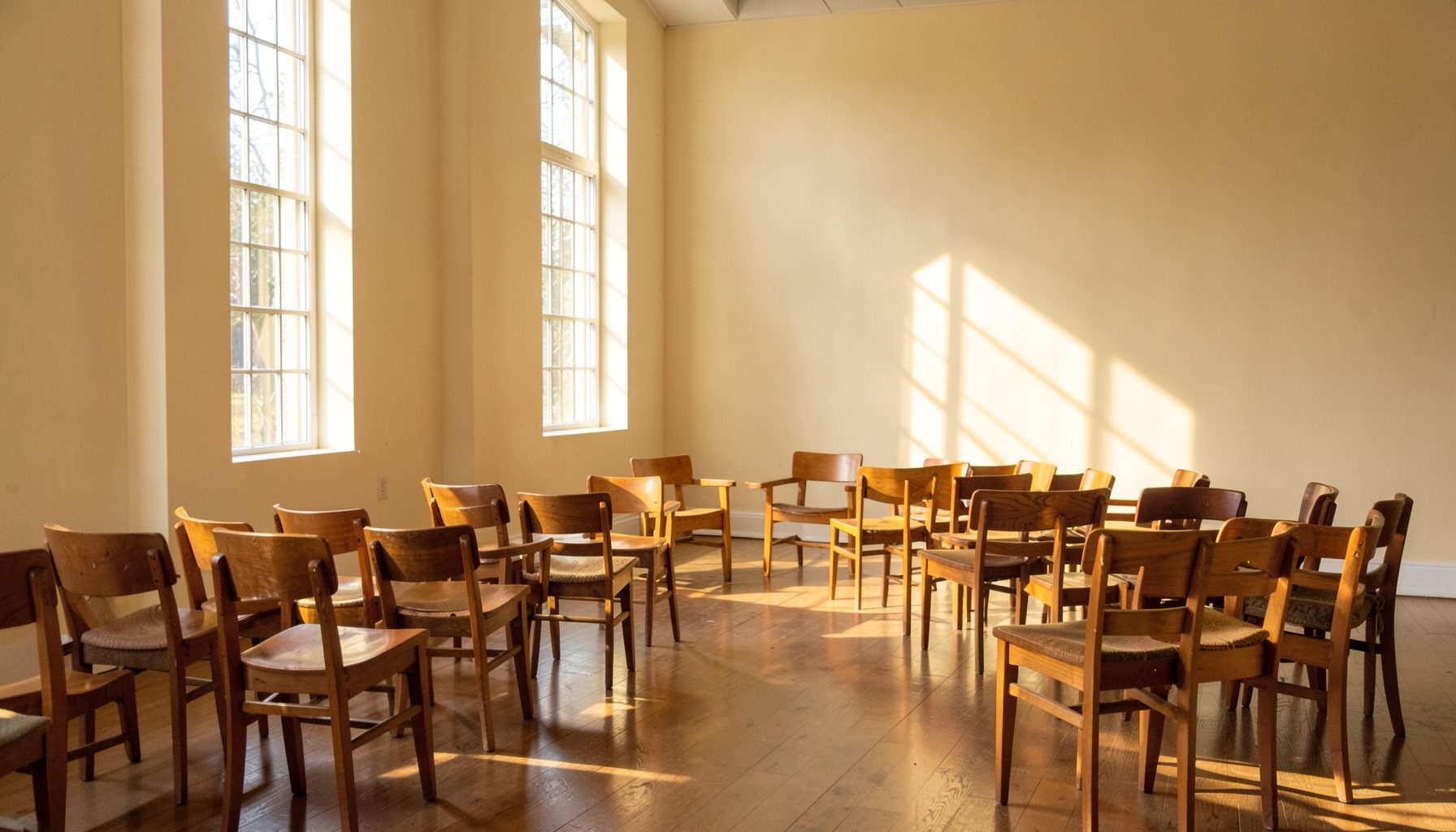 Warm sunlight filtering through tall windows into a quiet church fellowship hall, with soft cream walls and wooden chairs arranged in small circles