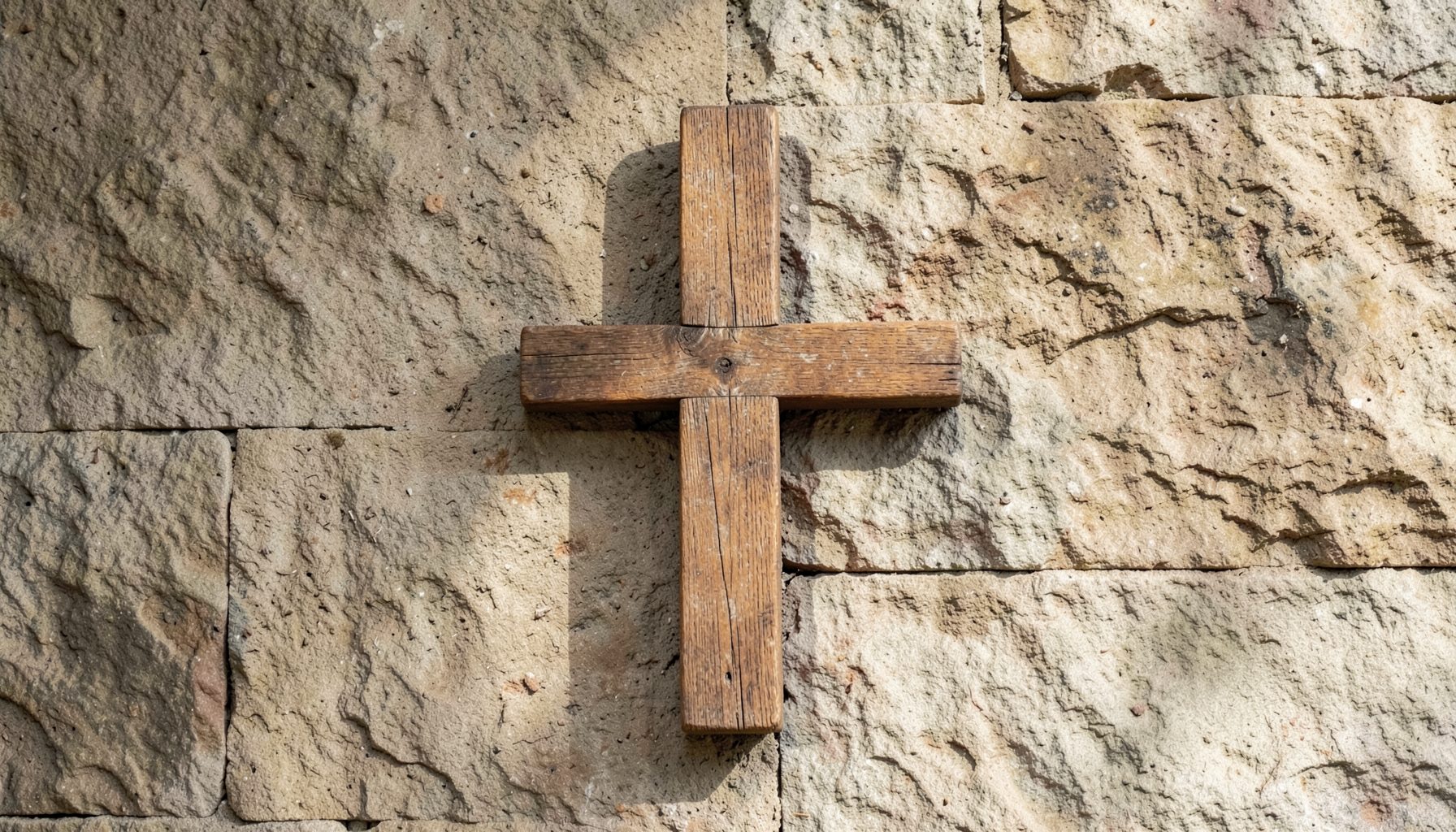 A wooden cross on a textured stone wall, softly lit by natural light with gentle shadows, in muted earth tones