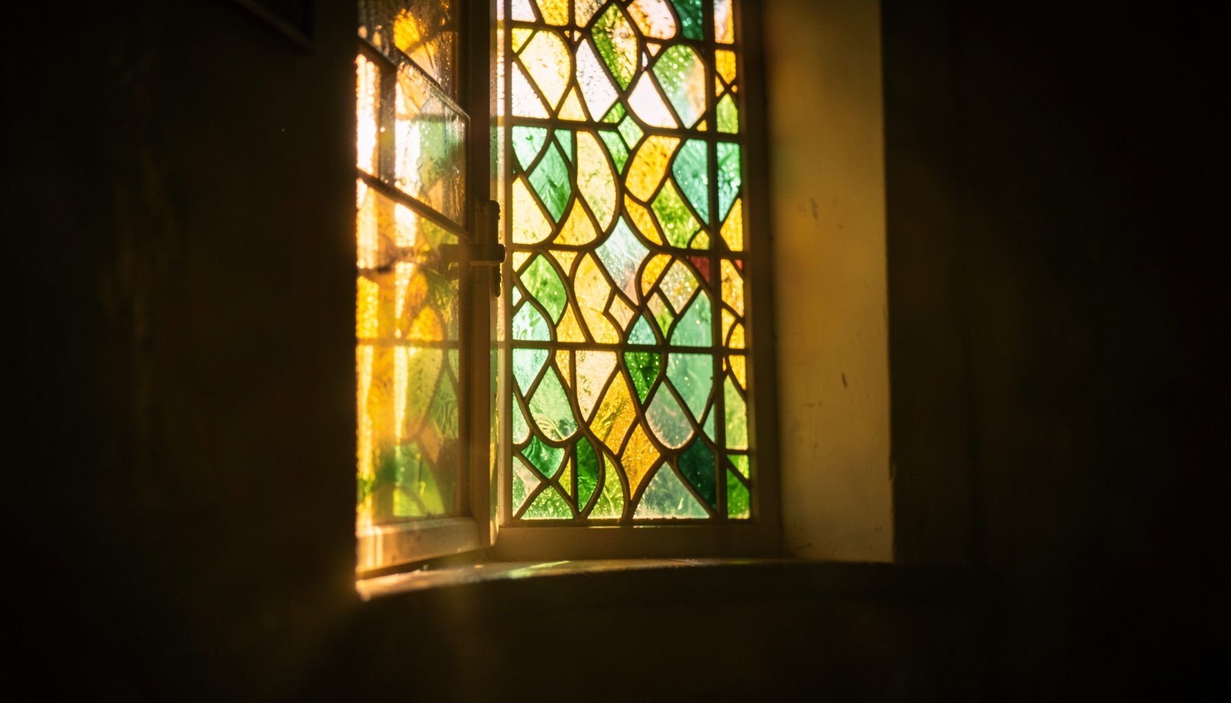 Warm sunlight filtering through a stained-glass window in soft amber, gold, and deep green tones, casting a peaceful glow